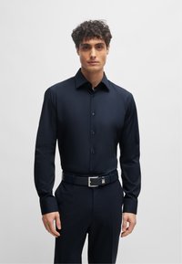 Camicetta da uomo a maniche lunghe di colore blu scuro con colletto a punta, bottoni frontali e cintura coordinata con fibbia in metallo. Tessuto liscio e aderente.