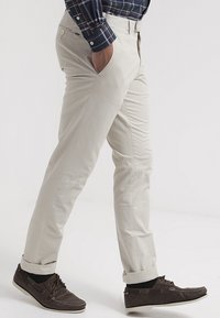Beige chinos med slim fit, sidofickor och kontrasterande sömmar. Kombineras med mörkbruna seglarskor, vilket visar en avslappnad stil.