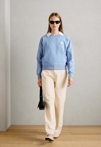 Sweatshirt bleu clair avec un col blanc et un logo, associé à un pantalon large de couleur crème. Le mannequin porte des lunettes de soleil noires et tient un petit sac.
