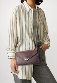 Brun handväska i konstläder med krokodilprägling, guldkedjestrap, och trekantigt lock, hålls mot en randig skjorta.