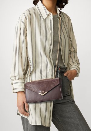 Bruine handtas van imitatieleren met croc-structuur, gouden kettingriem en driehoekige klepsluiting, vastgehouden tegen een gestreept shirt.