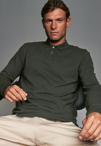 Polo de manga larga verde oscuro con dos botones, puños acanalados y un sutil detalle de logo. Combinado con pantalones de corte relajado en tonos claros.