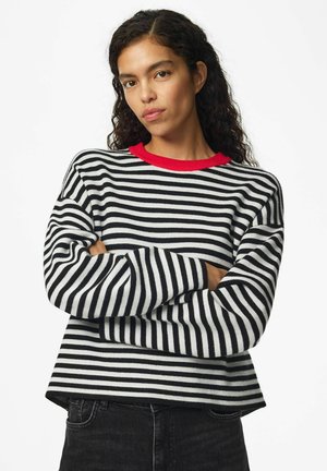 Femme aux cheveux bouclés foncés portant un pull rayé noir et blanc avec un col rouge, debout les bras croisés devant un fond blanc.