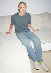 Grijze ronde hals t-shirt, lichtblauwe spijkerbroek en beige geweven instapschoenen met groene accenten. Zittend op een gestructureerde witte bank.