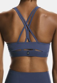 Sostenedor deportivo azul con un diseño de espalda cruzada, textura suave y etiqueta de logo. La tela parece elástica y transpirable, con detalles recortados.