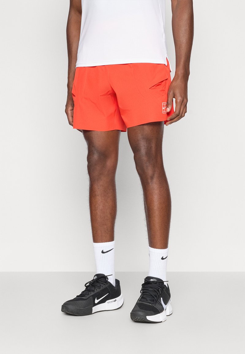 Φωτεινά πορτοκαλί αθλητικά σορτς με τσέπες, άνετη εφαρμογή και λεπτομέρεια με λογότυπο. Φοριούνται με μαύρα αθλητικά παπούτσια και λευκές κάλτσες.