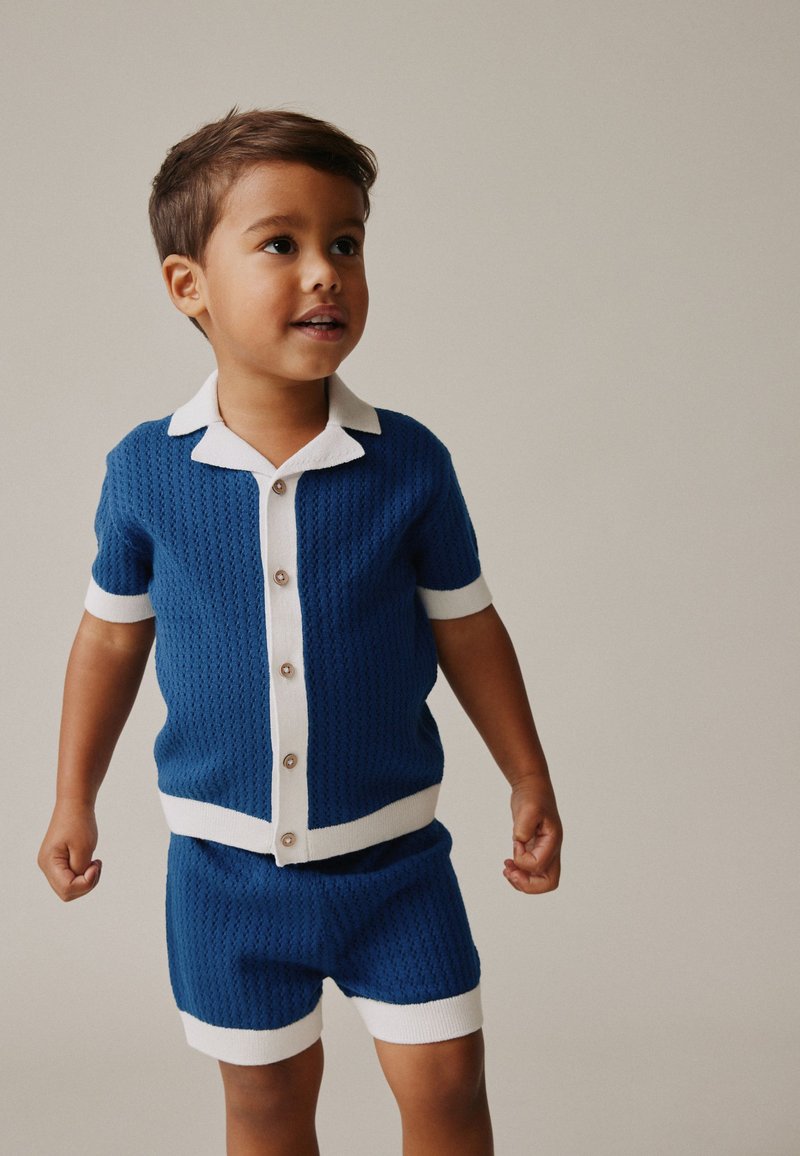 Ragazzo giovane che indossa una camicia a maniche corte con bottoni in maglia blu e pantaloncini abbinati con bordo bianco, in piedi davanti a uno sfondo semplice.