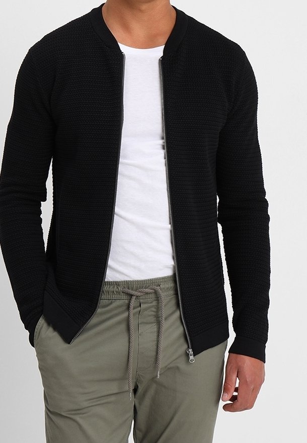 Veste bomber texturée noire avec une fermeture éclair à l'avant, des poignets côtelés et un col rond, associée à un t-shirt blanc et à un pantalon kaki.