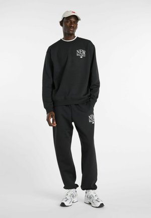 Zwarte New Balance trui en joggingbroek gedragen door een staande man met een witte pet en witte New Balance sneakers tegen een effen achtergrond.