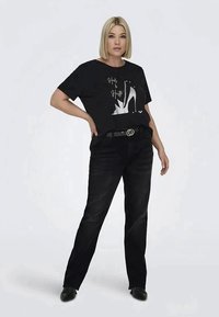 Juodas grafikų T-shirt su baltu aukšto kulno dizainu, derinamas su juodais džinsais ir plačiu diržu su apvalia sagtimi. Modelis stovi pasitikintis.