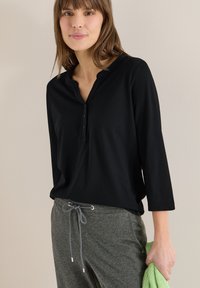 Jeune femme portant un t-shirt Henley noir à manches longues et un pantalon gris à cordon, tenant un tissu tricoté vert dans sa main droite.