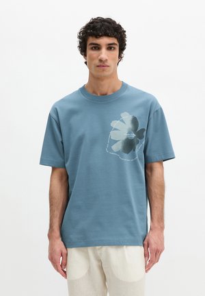 Mężczyzna o kręconych włosach, ubrany w niebieską koszulkę z dużym kwiatowym nadrukiem i białym napisem na piersi, w połączeniu z jasnymi spodniami.