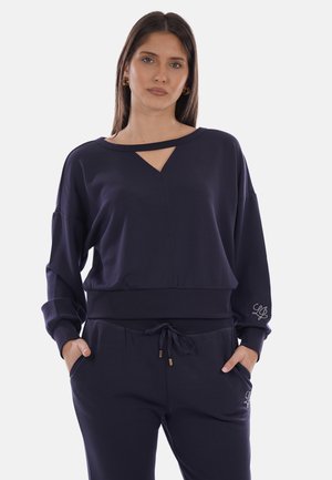 Donna che indossa una maglia a maniche lunghe blu navy con scollo a taglio e pantaloni abbinati con coulisse, mani nelle tasche, espressione neutra.