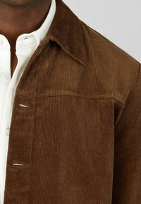 Bruine corduroy jas met een kraag, voorzien van een verticale ribbelstructuur en een enkele borstzak. Witte overhemden zichtbaar eronder.