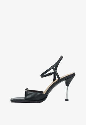 Czarna skórzana sandał z paskiem wokół kostki, odkrytymi palcami i metalicznym wysokim obcasem. Charakteryzuje się płaskim, kwadratowym czubkiem oraz minimalistycznymi detalami.