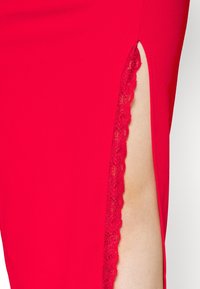 Robe rouge avec une fente haute sur le côté bordée de dentelle laissant entrevoir une partie de la jambe nue à droite.