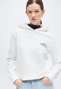 Femme aux cheveux bruns attachés en queue de cheval basse, portant un sweat à capuche blanc GANT, un jean bleu clair et de petites boucles d'oreilles créoles, les mains dans les poches, fond neutre.