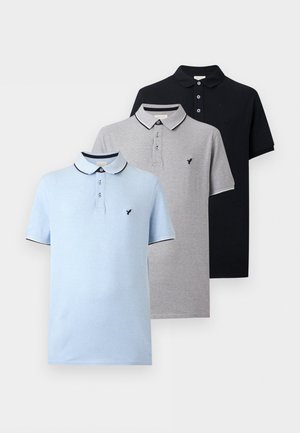 Homme portant un polo noir, un pantalon de jogging gris clair et des baskets blanches avec des bandes noires, debout sur un fond blanc.