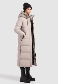 Dlhý svetlo béžový páperový kabát s kapucňou, horizontálnym prešívaním, zipsovými vreckami a čiernymi kovovými doplnkami, nosený cez čierne oblečenie a topánky.