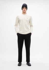 Cămașă bej deschis cu mâneci lungi, având un mic logo negru, purtată cu pantaloni negri și cizme negre. Modelul poartă o căciulă dark.
