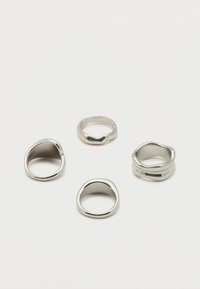 Quatre bagues en argent de forme irrégulière présentées sur une surface blanche unie, mettant en valeur des finitions lisses et polies.