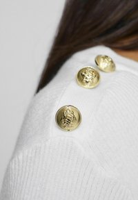 Witte gebreide trui met drie gouden sierknopen met reliëfdetails op de schouder, textiel met een geribbeld patroon.