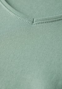 Detailní záběr na látku v mentolově zelené barvě s hladkou strukturou a designem s výstřihem do V. Viditelné šití podél výstřihu a lemování.