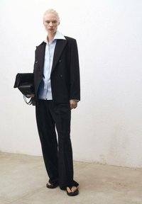 Černý dvouřadý blazer über světle modrou pruhovanou košili, doplněný černými širokými kalhotami a černými sandály, držící černou kabelku.