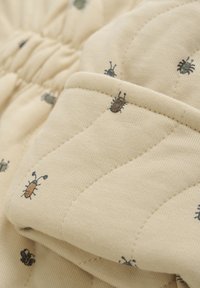Tissu beige matelassé avec de petits motifs d'insectes en vert et marron. Les détails comprennent un décolleté froncé et une couture texturée.