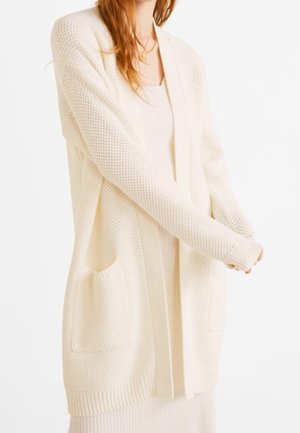 Frau trägt eine lange, cremefarbene Strickjacke mit strukturierten Ärmeln und großen vorderen Taschen über einem gerippten Kleid.