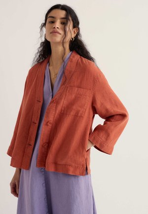 Göndör sötét hajú nő laza, rozsdavörös, gombos dzsekit visel, amely alatt levendulalila gombos ruha van, miközben egy egyszerű világos háttér előtt áll.