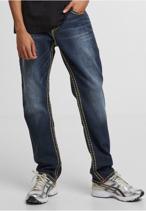 Dark blue jeans with contrasting yellow stitching, straight cut, and two side pockets. Worn with silver athletic shoes.