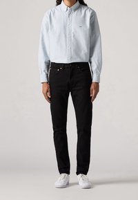 Person som står i en blå och vit randig skjorta med knappar, svarta jeans och vita sneakers mot en enfärgad bakgrund.