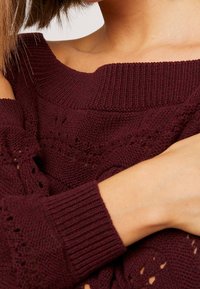 Bordowy sweter dzianinowy z dekoltem na ramieniu, teksturowanym wzorem, ściągaczami przy mankietach i ażurowymi detalami, ukazujący miękki materiał.