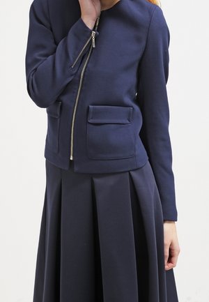 Femme portant une veste zippée bleu marine avec des poches sur le devant et une jupe plissée bleu marine sur fond blanc.
