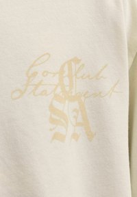 Tissu de couleur crème avec un texte décoratif beige pâle et des lettres stylisées "S" et "A" qui se chevauchent au centre.