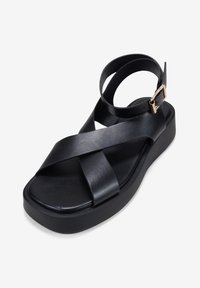 Black leather sandal with a cross strap design and adjustable ankle strap. Features a chunky platform sole and a metallic buckle accent.