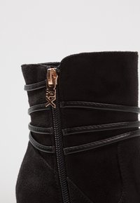 Stivale alla caviglia nero in suede con un design a fascia nera lucida. Presenta una cerniera laterale visibile con una linguetta in oro rosa.