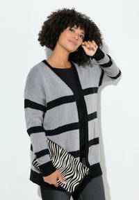 Harmaa ja mustaraidallinen kardigan, jossa on löysä istuvuus, neulepinta ja avoin etuosa. Yhdistetty mustaan puseroon ja seeprahmittaiseen laukkuun.