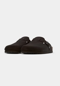 Σκούρα καφέ παπούτσια τύπου clogs χωρίς κορδόνια από σουέντ με λείο εξωτερικ ό, στρογγυλεμένη μύτη, ρυθμιζόμενο λουράκι και υφασμένο καουτσούκ στη σόλα.