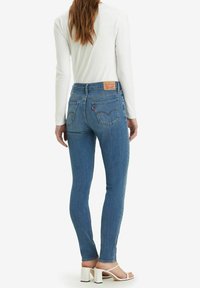 Kapein farkut keskisinisessä denimissä, joissa on takataskut ja merkkilaatta. Yhdistetty valkoiseen pitkähihaiseen puseroon ja valkoisiin korkokenkiin.