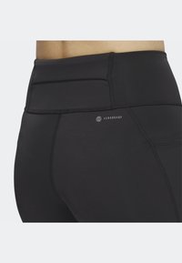 Zbliżenie na osobę, która nosi czarne legginsy sportowe z wysokim stanem, z logo "AEROREADY" na tylnym pasie.