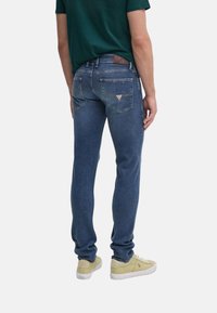 Jeans de mezclilla ajustados en azul oscuro, con un parche de logo de cuero, dos bolsillos traseros con costuras decorativas y una textura suave.