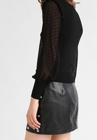 Schwarzer Pullover mit strukturierten, transparenten Ärmeln und Rüschensaum, kombiniert mit einem hoch taillierten schwarzen Lederrock mit silbernen Knöpfen.