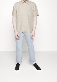 Beige linnen shirt met korte mouwen en een borstzak, gecombineerd met lichtblauwe rechtgesneden jeans en zwarte sneakers met witte zolen.