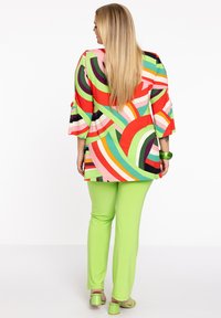 Blouse à motifs colorés avec des manches larges, ornée de tourbillons rouges, verts et roses. Associée à un pantalon vert ajusté et des sandales assorties.