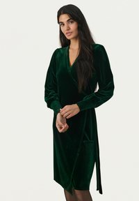Grünes Samt-Wickelkleid mit langen Puffärmeln, tiefem V-Ausschnitt, einer Seitentasche und einer Bindeschließe. Glatte Textur und eleganter Schnitt.