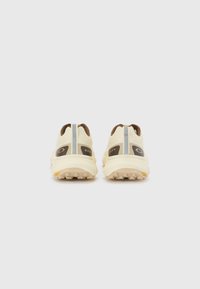 Helle beige Sportschuhe mit einem strukturierten Obermaterial, braunen Akzenten und einem markanten Logo auf der Rückseite. Verfügt über eine robuste Sohle für besseren Halt.