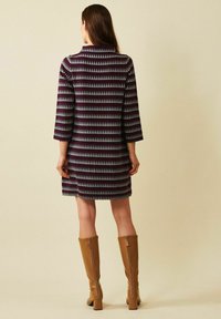 Kvinna som bär en knäl�ång, randig klänning med små trämönster och trekvartslånga ärmar, i kombination med beige knähöga klackstövlar, stående.