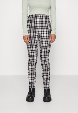 Osoba nosząca czarno-białe spodnie w kratę, jasnoszary długi crop top z długim rękawem oraz czarne masywne buty, stoi na jednolitym jasnym tle.
